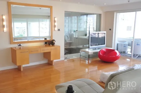 Condominium for rent in Khlong Toei, Bangkok - Bright living area with wooden floors, floating vanity and mirror, glass‑walled bathroom, TV unit and balcony access.