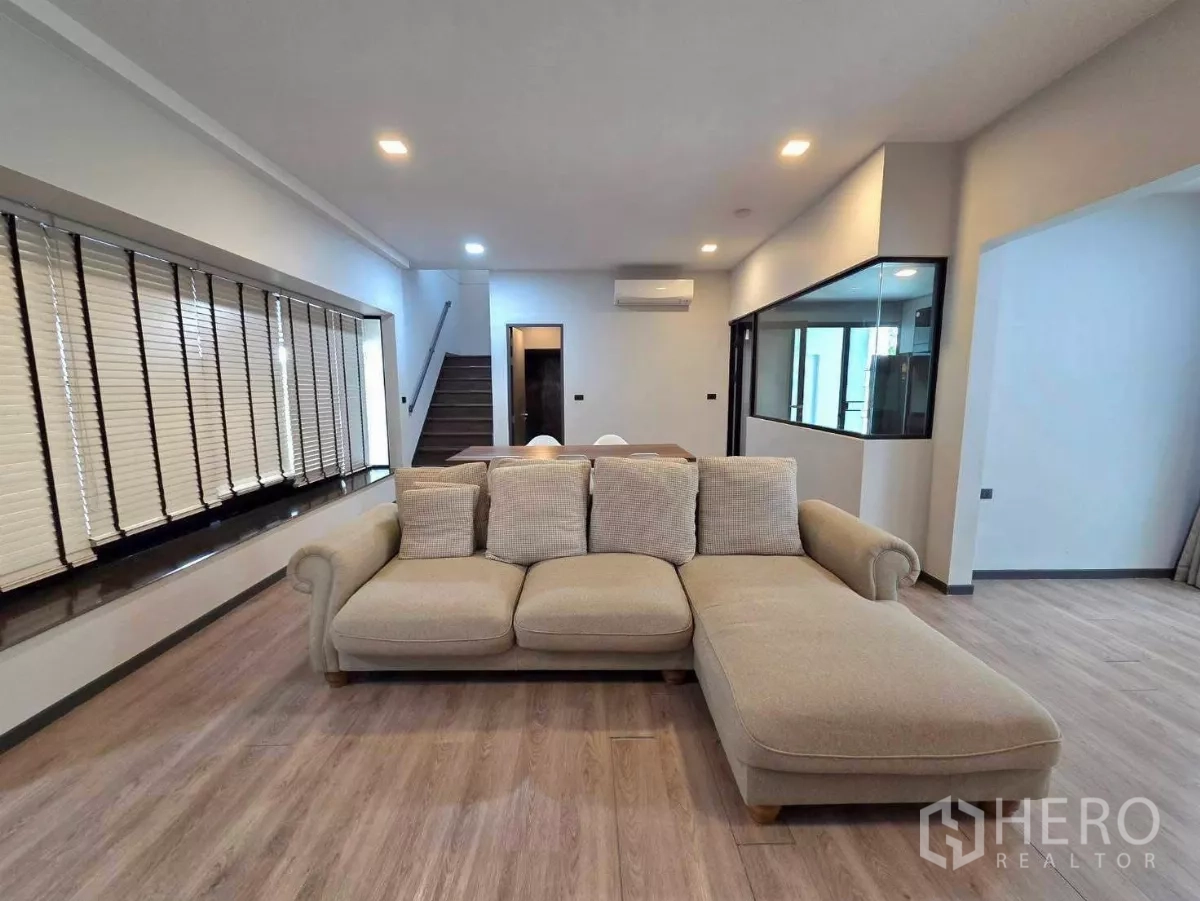Detached House for rent in Bang Phli, Samut Prakan - Bright living room with L-shaped sofa, wood floors, staircase and a glass-partitioned kitchen.