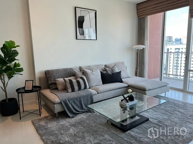 Condominium for rent in Watthana, Bangkok - Cozy lounge corner with a gray L-shaped sofa, glass coffee table, large rug, and balcony access.