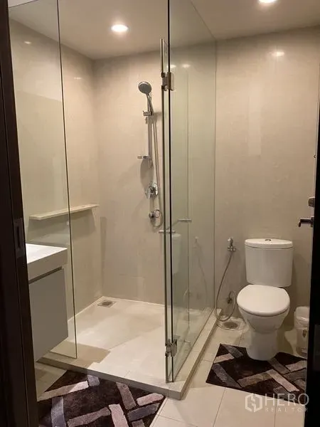 Condominium for rent in Watthana, Bangkok - Bathroom with glass‑enclosed shower, toilet and recessed ceiling lights on beige tiles.