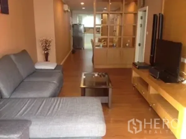 Condominium for rent or sale in Watthana, Bangkok - Living room with gray sectional sofa, glass coffee table and TV console on wood floor.