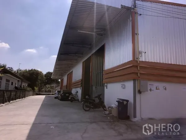 Warehouse for rent in Lam Luk Ka, Pathum Thani - Corrugated-metal warehouse exterior in Lam Luk Ka with high roof, large roll-up doors, covered loading bay and wide concrete driveway for truck access.
