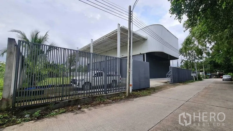Warehouse for rent in Mueang Nonthaburi, Nonthaburi - Corner view of the warehouse compound with perimeter fencing, canopy roof and greenery along the road.