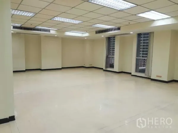 办公空间 出租在Ratchathewi, Bangkok - 明亮的开放式办公室，网格吊顶灯、米色墙面与高窗，可见周边建筑。