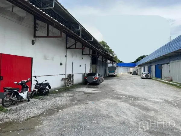 货栈 出租在Mueang Samut Prakan, Samut Prakan - 仓库园区内宽阔碎石车道，红色装卸门，汽车与摩托车停车位