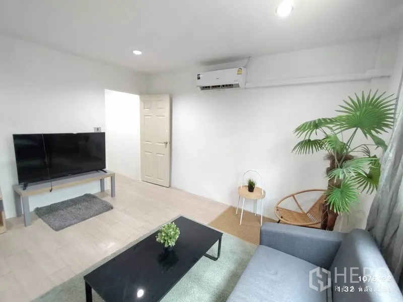 Home Office for rent - Living room view showing TV on stand, wall-mounted air conditioner, side tables and a potted palm by the window.