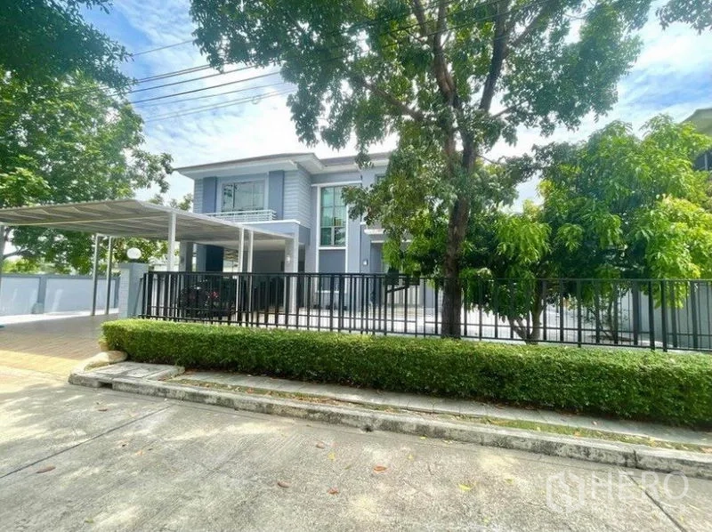 独立式住宅 出租在Prawet, Bangkok - 街景角落地块，黑色围栏、成树与宽阔车道。