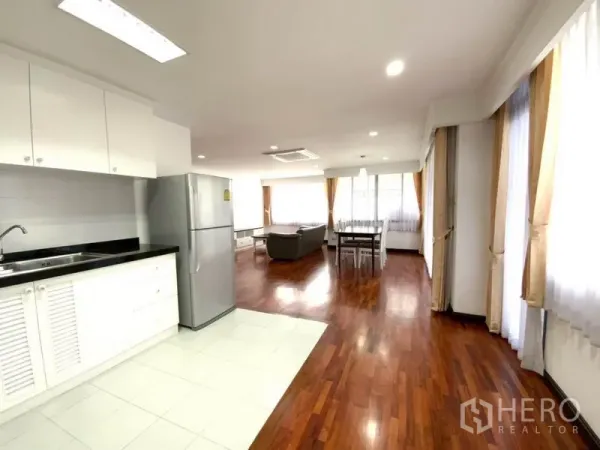 公寓 出租在Watthana, Bangkok - 开放式厨房配白色橱柜与不锈钢冰箱，连接宽敞的客餐区，木地板与落地窗帘环绕。