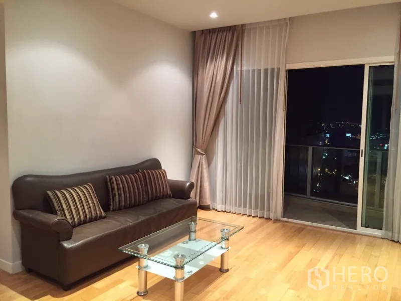 Condominium for rent in Khlong Toei, Bangkok - Bright living room with leather sofa, glass coffee table, wood floors, and balcony city view at night.