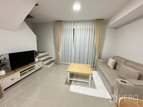 Townhouse for rent in Bang Phli, Samut Prakan - Bright living room with beige sofa, TV console, stairs and floor‑to‑ceiling curtains.