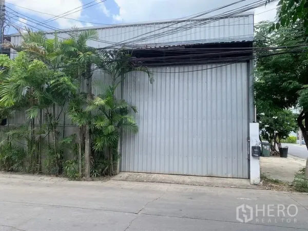 คลังสินค้า ให้เช่าในBang Kapi, Bangkok - ภาพระยะใกล้ประตูเมทัลชีทขนาดใหญ่ พร้อมต้นปาล์มข้างทางเข้าโกดัง