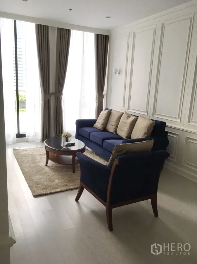 Condominium for rent in Pathum Wan, Bangkok - Bright living room with navy sofa set, paneled walls, and floor-to-ceiling curtains.