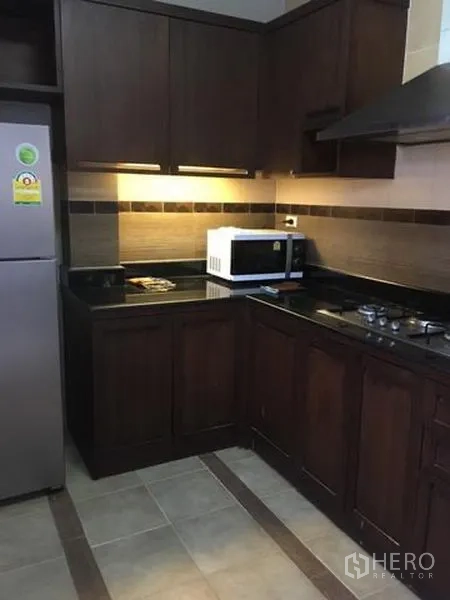 Detached House for rent in Bang Kapi, Bangkok - L-shaped kitchen with dark wood cabinets, granite counters, gas hob, hood, microwave and fridge.