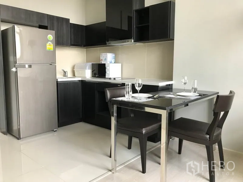 Condominium for rent in Khlong Toei, Bangkok - Modern kitchenette with dark cabinetry, stainless refrigerator, and a two-seat dining table set.