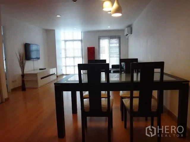Condominium for rent in Bang Rak, Bangkok - Dining area facing a bright living room with wall-mounted TV, large windows, and a glass dining table on wooden floors.