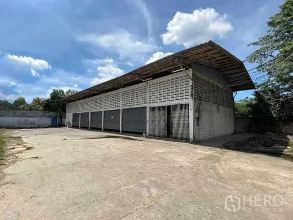 Warehouse for rent in Bang Na, Bangkok - Single-storey warehouse with multiple roller shutters and a wide concrete yard under a blue sky in Bang Na, Bangkok.