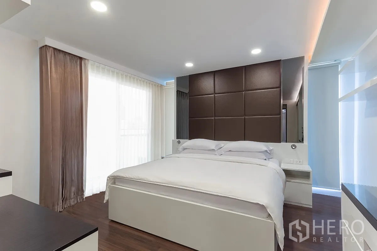 Condominium for rent in Khlong Toei, Bangkok - Second bedroom with padded headboard, sheer curtains, wood floors, and soft lighting.