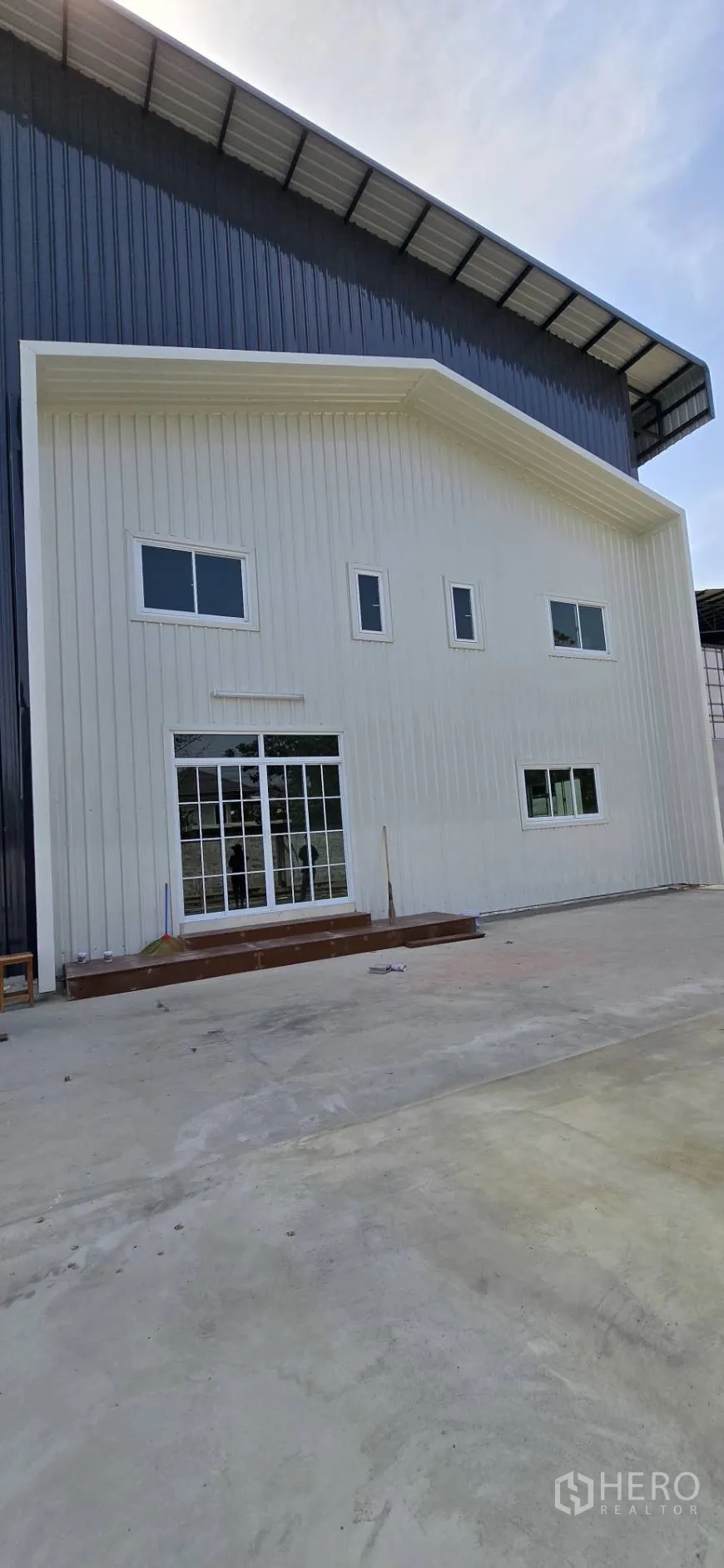 โรงงาน for rent in Mueang Samut Sakhon, Samut Sakhon - ด้านหน้าส่วนออฟฟิศสีขาวติดกับตัวโกดัง พร้อมประตูกระจกบานเลื่อนและหน้าต่างหลายบาน.