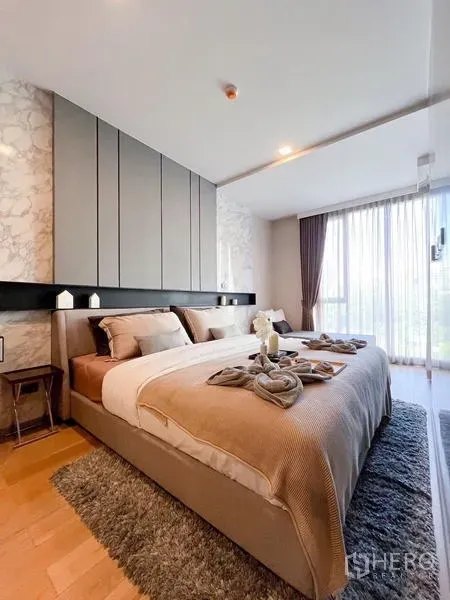 Condominium for rent in Watthana, Bangkok - Primary bedroom with king-size bed, warm neutral tones, wood flooring, and floor-to-ceiling window.
