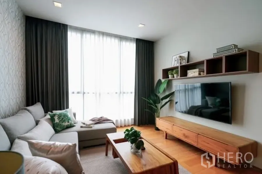 公寓 出租在Watthana, Bangkok - 温馨客厅，L 型沙发，大屏电视，壁挂置物架与落地窗。