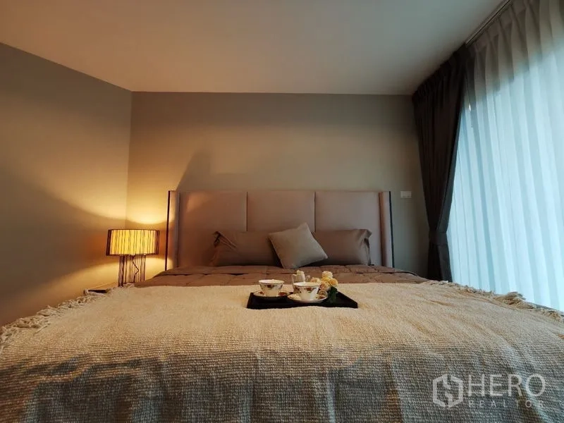 Condominium for rent or sale in Huai Khwang, Bangkok - Master bed with padded headboard beside full-height curtains, tray with tea on the blanket.
