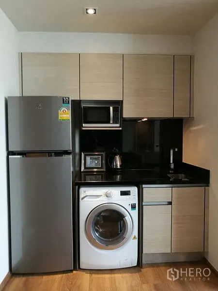 Condominium for sale in Khlong Toei, Bangkok - Compact kitchenette with fridge, microwave, washing machine and black stone countertop.