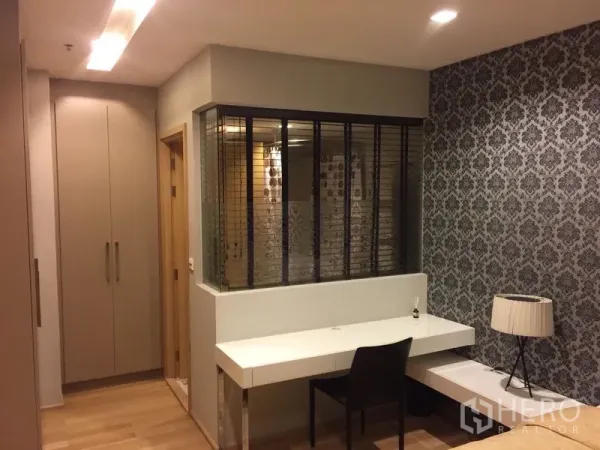 Condominium for rent in Khlong Toei, Bangkok - Work desk and built-in wardrobe beside a glass-partitioned room with patterned wallpaper and wooden floors.