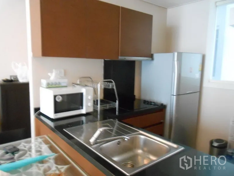 Condominium for rent in Watthana, Bangkok - Compact modern kitchen with black granite countertop, stainless sink, microwave, and full-size fridge.