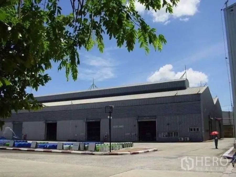 โรงงาน ขายในRayong - โรงงานโครงเหล็กผนังเมทัลชีท หลังคาทรงจั่วสูง มีประตูม้วนหลายจุดและลานคอนกรีตกว้าง ใต้ท้องฟ้าโปร่ง มีต้นไม้ด้านหน้า