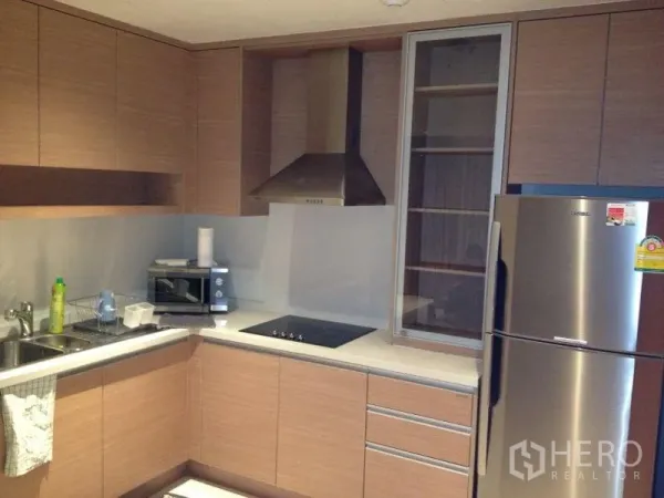 Condominium for rent or sale in Khlong Toei, Bangkok - Modern L-shaped kitchen with wood cabinetry, cooktop, range hood, microwave, and stainless refrigerator.