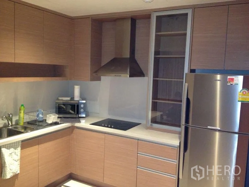 Condominium for rent or sale in Khlong Toei, Bangkok - Modern L-shaped kitchen with wood cabinetry, cooktop, range hood, microwave, and stainless refrigerator.