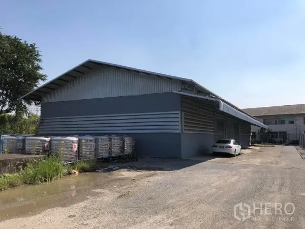 Factory for rent in Bang Bua Thong, Nonthaburi - Exterior view of a gray metal factory with covered loading area and parking on a sunny day.
