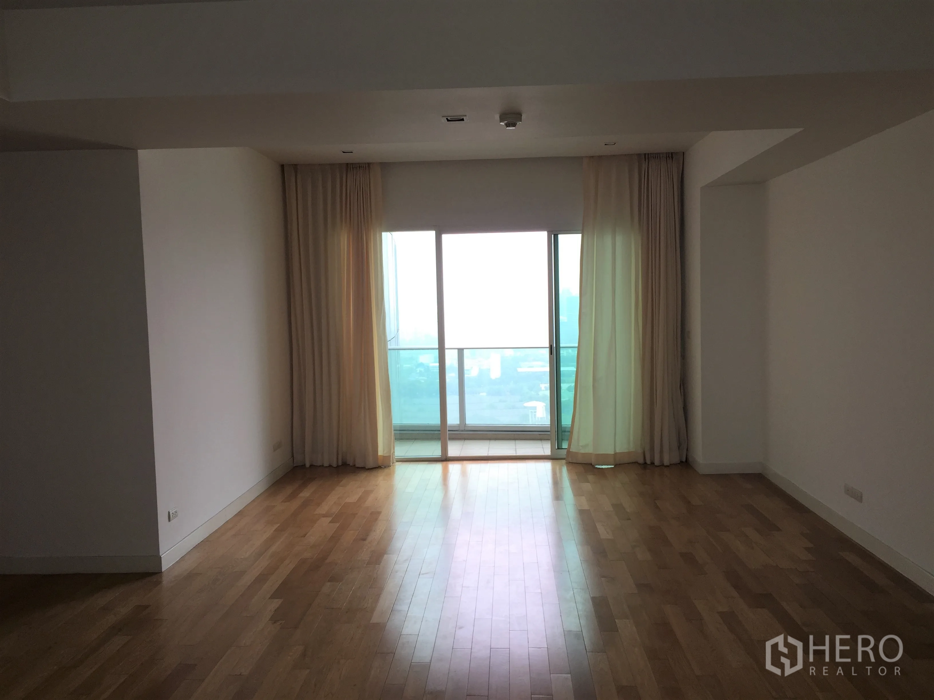 Condominium for rent in Khlong Toei, Bangkok - Spacious living room with hardwood floors, sheer curtains and a sliding door opening to a balcony with Bangkok skyline views.