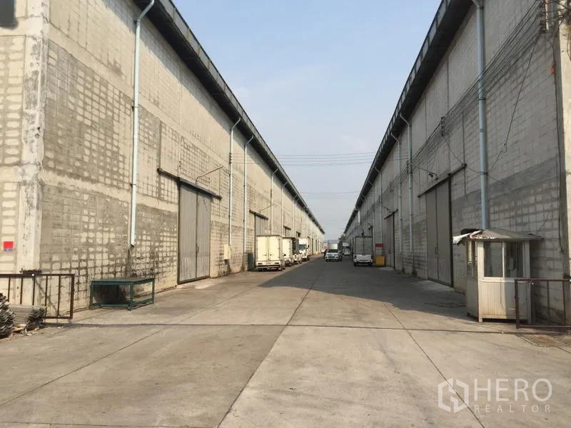 Warehouse for rent in Mueang Pathum Thani, Pathum Thani - Long service road between warehouse rows with trucks and a small guard booth.