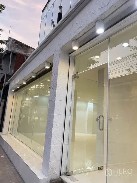 店屋 出租在Dusit, Bangkok - 白色商铺外立面，整面玻璃与射灯，傍晚街景，曼谷Dusit区。