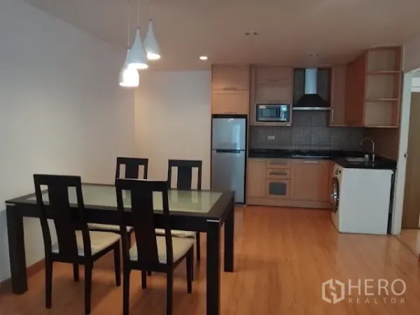 Condominium for rent in Bang Rak, Bangkok - Open-plan dining and kitchen with a glass-top table, pendant lights, built-in cabinetry, fridge, microwave, and washing machine.