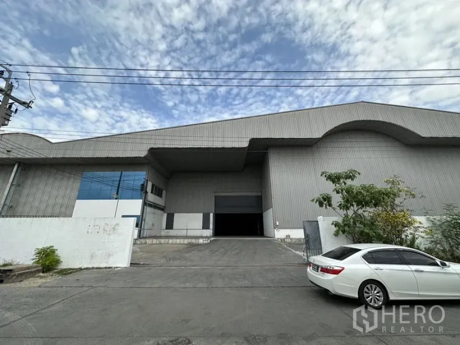 Warehouse for rent in Mueang Samut Prakan, Samut Prakan - Front view of a large metal-clad warehouse with ramped driveway, roll-up loading door and canopy, with parking in front.