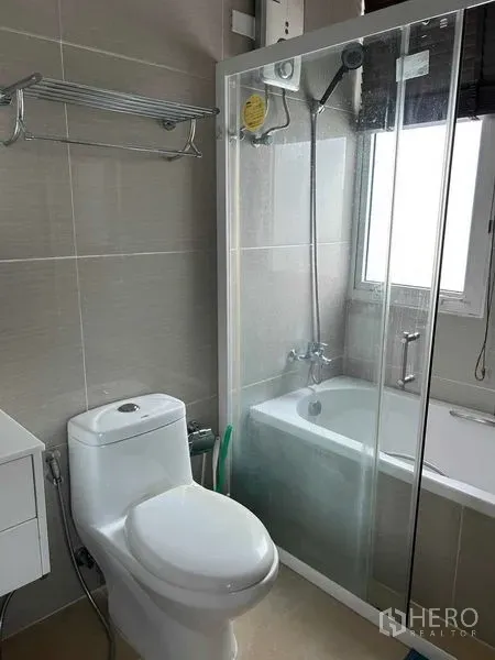 Condominium for sale in Huai Khwang, Bangkok - Modern bathroom with toilet, glass shower over bathtub and window.