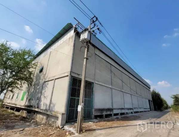 Warehouse for rent or sale in Sai Noi, Nonthaburi - Corner exterior of a large concrete warehouse with green roof, power lines, and a side access road in Sai Noi.