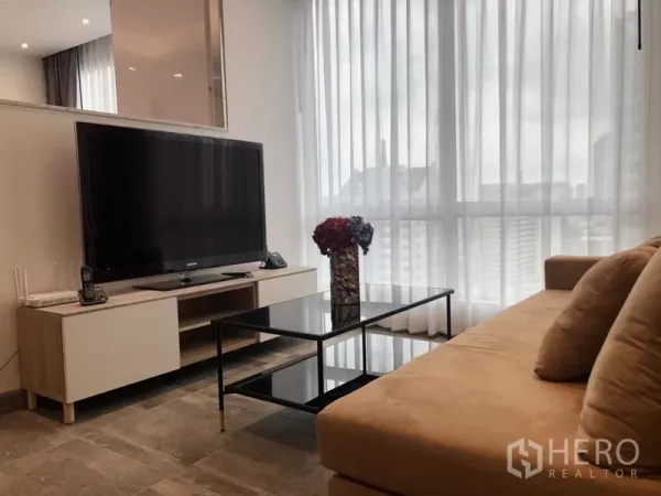 คอนโด ให้เช่าในSathon, Bangkok - ห้องนั่งเล่นสว่าง ทีวีจอใหญ่บนตู้วางทีวี โต๊ะกลางกระจก และผ้าม่านโปร่งสูงจรดเพดานมองเห็นวิวเมือง