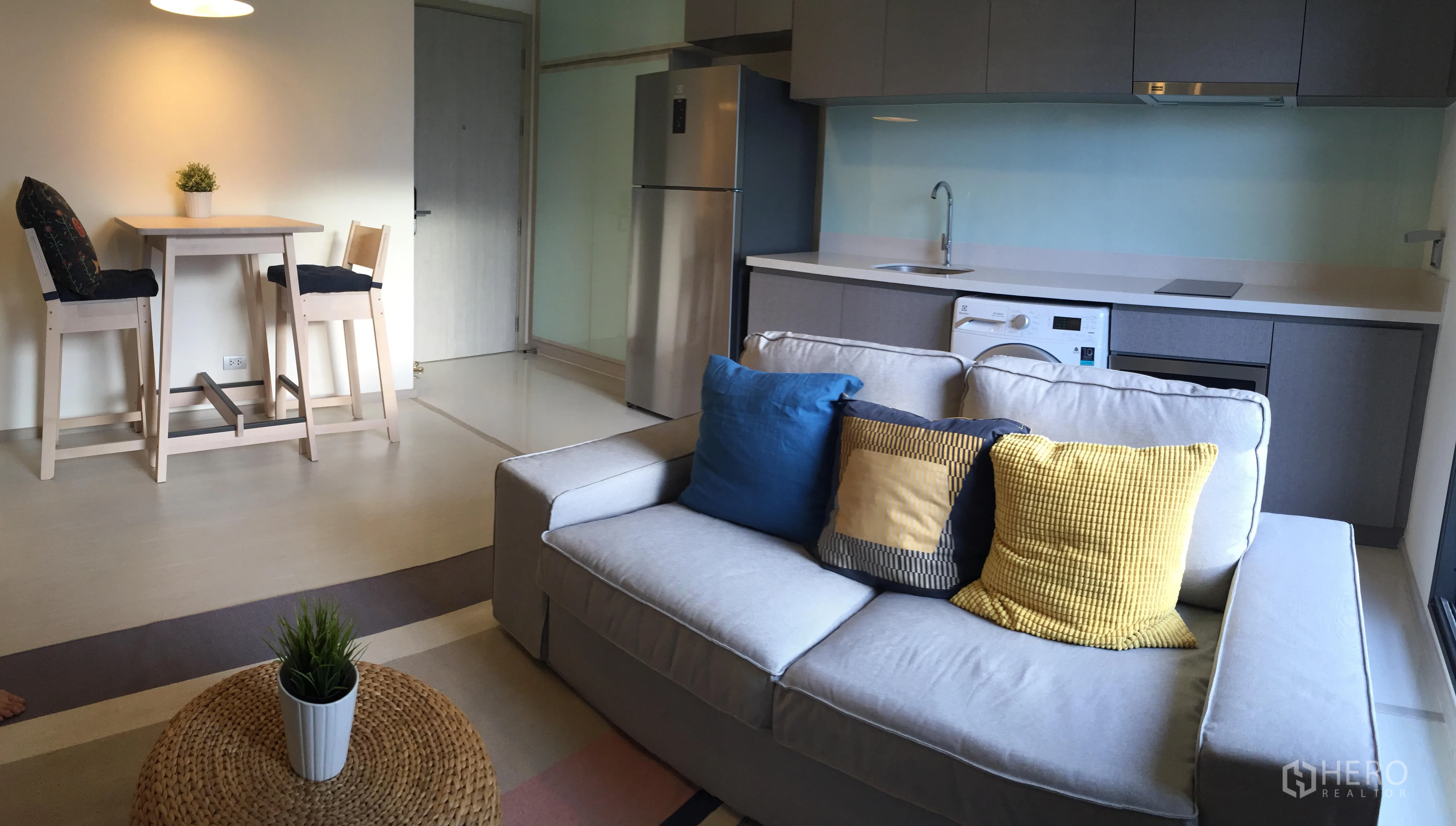 Condominium for rent in Khlong Toei, Bangkok - Open-plan living room with a gray sofa, colorful cushions, a bar-height dining set and a modern kitchen with fridge and washer.