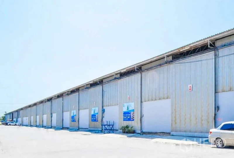 Warehouse for rent in Bang Sao Thong, Samut Prakan - Row of metal-clad warehouse units with multiple roller doors and a parking area under clear sky.