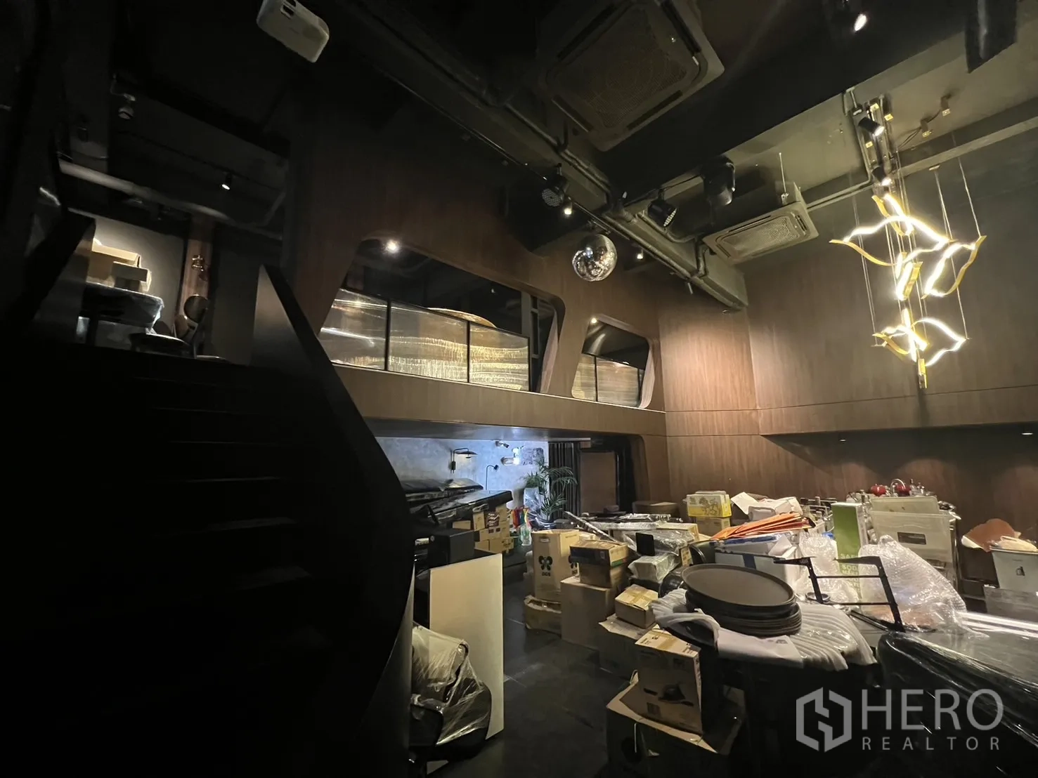 Shophouse for rent in Watthana, Bangkok - Open-plan level with high ceiling, staircase and stacked supplies under warm lighting.