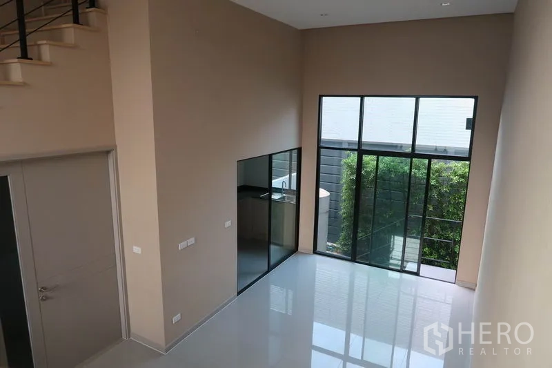 Townhouse for rent in Suan Luang, Bangkok - Bright living room with tall windows to a small garden and a glass-partitioned kitchen.