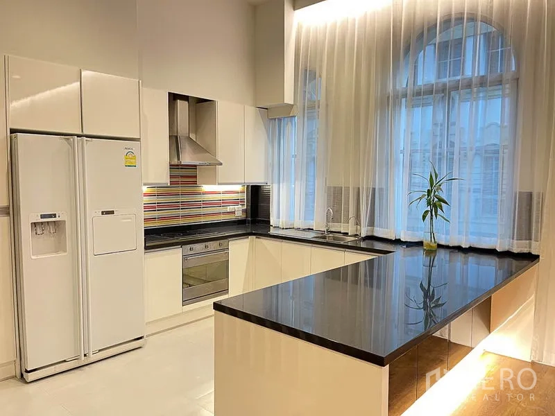 Townhouse for rent in Yan Nawa, Bangkok - Modern white L-shaped kitchen with black granite countertops, range hood and double-door refrigerator beside tall curtained windows.
