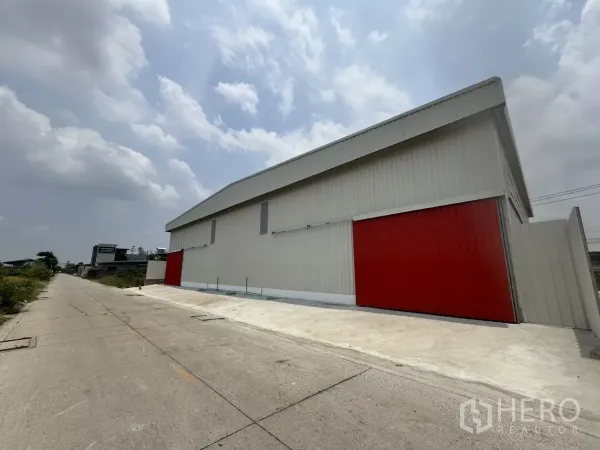Warehouse for rent in Lam Luk Ka, Pathum Thani - Side elevation of the warehouse showing long façade, red doors and a wide concrete driveway for truck access.