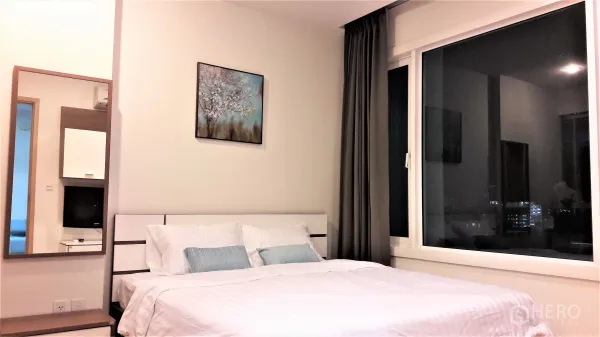 Condominium for rent in Khlong Toei, Bangkok - Bright bedroom with large window, city night view, artwork above a white bed and blackout curtains.