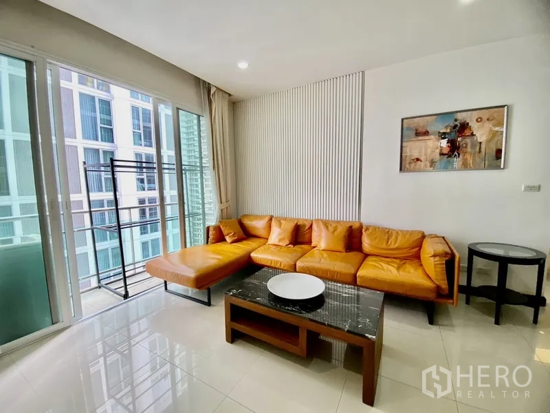 公寓 出租在Watthana, Bangkok - 采光充足的客厅，橙色皮沙发与石面茶几，推拉门通往阳台，可见周边高楼景观。