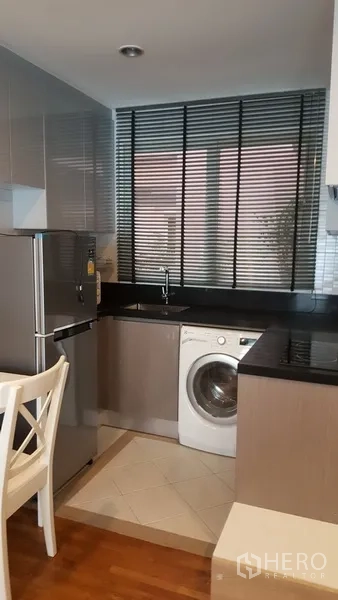 Condominium for rent in Watthana, Bangkok - Compact kitchen with black countertops, window blinds, stainless refrigerator, and a front-load washing machine.