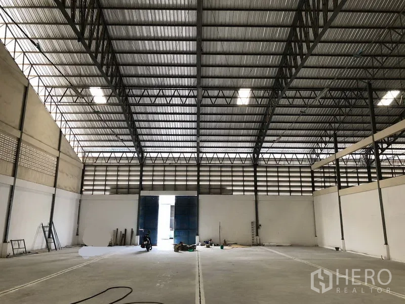 Warehouse for rent in Bang Phli, Samut Prakan - Spacious warehouse interior featuring a high steel‑truss roof, skylights and a smooth concrete floor leading to wide blue doors.